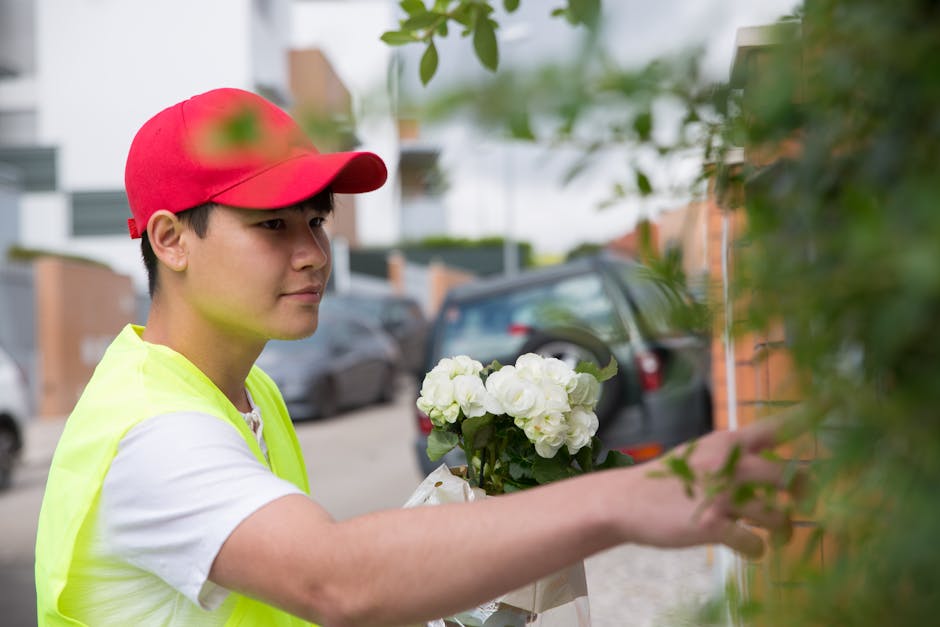 A young delivery person wearing a red cap and a high-visibility yellow vest is placing a bouquet of fresh white flowers, likely roses and carnations, against a green leafy shrub near a brick wall. The